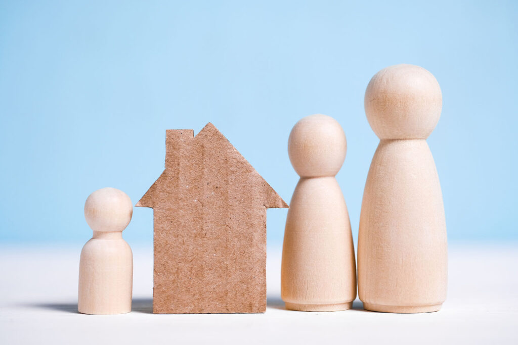 Vier Holzfiguren in Familiengröße stehen neben einem Haus aus Pappe – Sinnbild für ein Kind, das in seiner Familie zu Hause bleibt